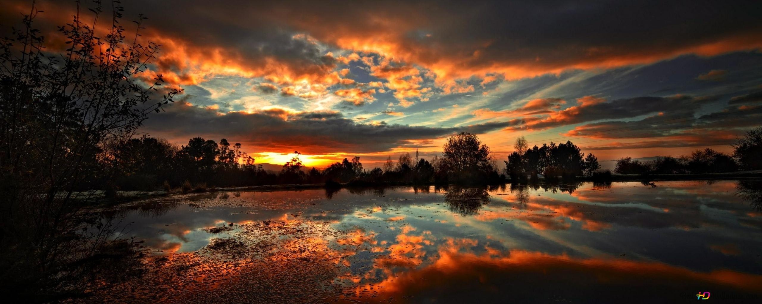 2560x1030 The reflection of the trees in the lake water with the sunlight that paints the sky red among the dark clouds 2K wallpaper download, Dual Screen